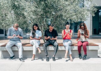Group of diverse adults sitting outdoors using smartphones under a tree.