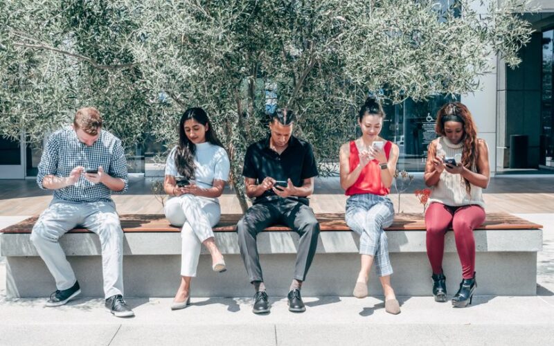 Group of diverse adults sitting outdoors using smartphones under a tree.